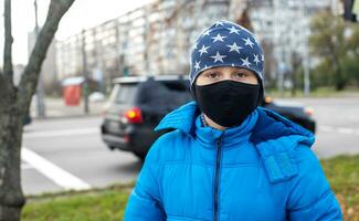 boy in face mask on street near road with cars. Child walks during quarantine and lockdown. child with flu, influenza, pollution in bad epidemic situation, among patients with coronavirus photo