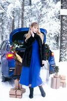 Woman in winter snowy forest in blue dress next to blue car decorated with Christmas decor. Christmas and winter holidays concept. photo