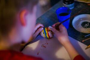 un niño decora un Pascua de Resurrección huevo en el colores de el arcoíris. un niño sostiene un huevo y pinturas eso con un cepillar. preparando para el celebracion de Pascua de Resurrección. foto