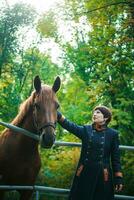 A woman in a blue long coat and with short dark brown hair extended her hand to stroke the horse. The horse pricked up his ears. A train for horses against the background of a green forest. photo