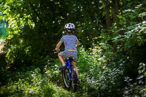 el pequeño chico en un a rayas traje va en un bosque la carretera. niño en un protector bicicleta casco montando un azul bicicleta. foto