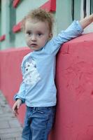 Cheerful little beautiful girl stands near a red wall. The child raised his hand walking down the street. photo