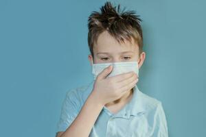 boy in medical mask on blue background. child coughs with hand covering mouth. Complications after viral infection. child with flu, influenza protected from viruses among patients with coronavirus photo