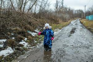 A child on a rural road with streams from melted snow in the spring. Happy toddler in rubber boots in gnawing and water in cloudy weather photo