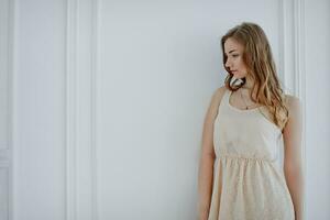 hermosa joven niña en beige vestir con largo rubio pelo soportes cerca blanco pared. niña esperando para regalo por marzo 8. joven mujer sin flores en internacional De las mujeres día. foto