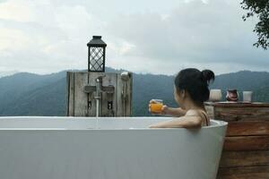 uno asiático mujer sin prisa relaja en un bañera y bebidas naranja jugo, descansando en panorámico montaña rango ver de tropical bosque, el tranquilo invisible paisaje natural viaje recurso para vacaciones. foto