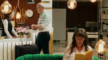 Caucasian man arriving at luxury hotel lobby, goes for check in registration at front desk. Senior traveller heading to front desk with luggage for room booking procedure, retirement age. video