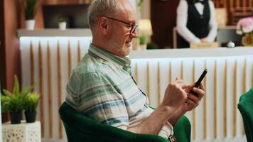 gepensioneerd Mens controle smartphone app in lounge Oppervlakte Bij vijf ster hotel, gebruik makend van online browser naar navigeren Aan sociaal media. senior reiziger ontspannende in vakantie terugtrekken lobby. handheld schot. video