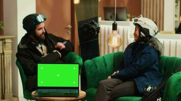 Couple with snow attire in hotel lobby engage in conversation, laptop running greenscreen display on table. Device showing isolated mockup template, diverse people talk about winter sport. video