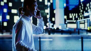 Asian man talking over the phone with wife using cellphone to feel less bored while strolling around dimly illuminated empty avenues at night, blurry cityscape background with bokeh lights photo