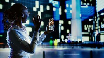 Woman greeting husband in online videocall while strolling around city streets at night. Person using cellphone to show friend urban surroundings in dimly illuminated metropolitan centre photo