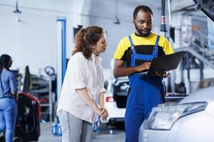Repairman at auto repair shop conducts yearly vehicle checkup, informing customer about needed engine mending. BIPOC garage worker discussing with customer after finishing car maintenance photo