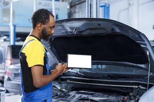 African american mechanic trainee uses mockup tablet in car service to watch tutorial on how to replace motor. Garage workplace intern looks up training videos online on isolated screen device photo