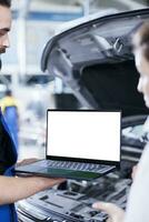 Licensed repairman in garage using mockup laptop to follow checklist while doing maintenance on car. Meticulous expert in auto repair shop does checkup on vehicle helped by isolated screen device photo