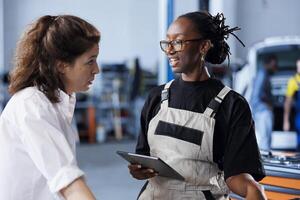 BIPOC mechanic in car service uses tablet to calculate invoice after fixing broken automobile engine. Trained auto repair shop worker uses device to inform woman of final costs after mending vehicle photo
