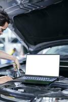 Mockup laptop on car with hood open while mechanic in blurry background replaces compressor belt. Isolated screen device next to skilled garage worker servicing client vehicle photo