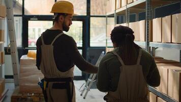 Meticulous employee walking through warehouse aisles scanning cardboard boxes on shelves under team leader guidance, using digital devices to update info before sending packages for shipping photo