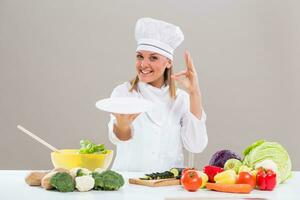 Portrait of cheerful female chef sitting at the table,showing empty plate and ok sign while making healthy meal. photo