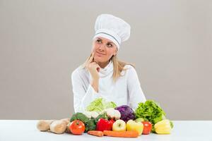 Cheerful female chef is sitting at the table with bunch of vegetable and thinking what to cook. photo