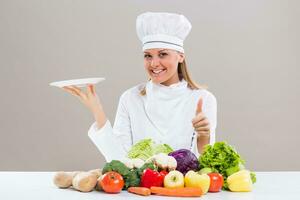 Female chef surrounded with vegetables holding plate showing thumb up photo