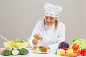 Cheerful female chef is sitting at the table with bunch of vegetable and decorating prepared meal. photo