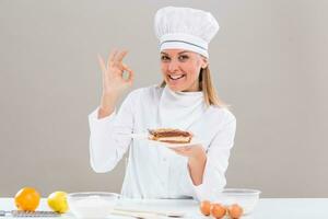 Beautiful female confectioner is showing slice of cake and ok sign  while sitting at the table photo