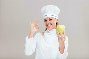 hermosa hembra cocinero es demostración Okay firmar y manzana en gris antecedentes. foto