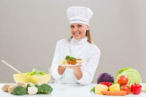 Content female chef is sitting at the table with bunch of vegetable showing prepared meal. photo