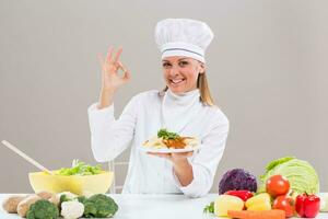 Cheerful female chef is sitting at the table with bunch of vegetable ,showing prepared meal and ok sign. photo