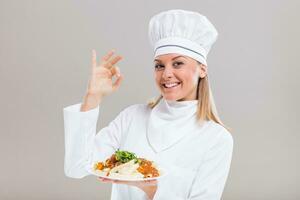 Beautiful female chef is showing ok sign and prepared meal on gray background. photo