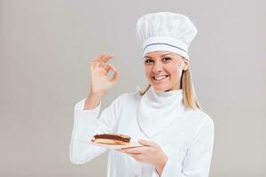 Beautiful female confectioner is showing slice of cake and ok sign on gray background. photo