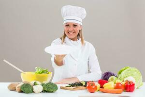 retrato de alegre hembra cocinero sentado a el mesa y demostración vacío plato mientras haciendo sano comida. foto