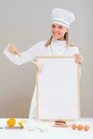 Beautiful female confectioner is showing whiteboard with slice of cake and ingredients for cake at the table. photo