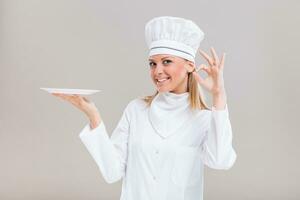 Beautiful female chef is holding plate and showing ok sign on gray background. photo