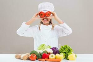 Cheerful female chef is covering her eyes with tomato while sitting at the table with bunch of vegetable. photo