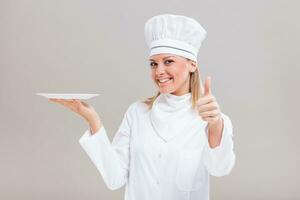 Beautiful female chef is holding plate and showing thumb up on gray background. photo