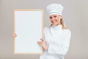 Portrait of beautiful female chef holding whiteboard on gray background. photo