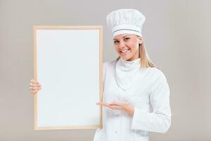 Portrait of beautiful female chef showing empty whiteboard on gray background. photo