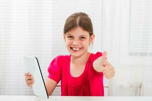 pequeño niña utilizando digital tableta y demostración pulgar arriba foto