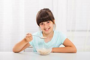 linda pequeño niña es sentado a el mesa y comiendo cereales para desayuno. foto