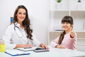 Female doctor and child sitting at the medical office and little girl showing thumb up photo