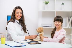 Female doctor and little girl examining teddy bear at medical office photo