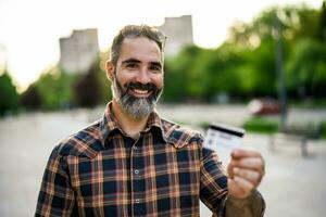 retrato de moderno empresario con barba participación crédito tarjeta mientras en pie en el ciudad calle foto