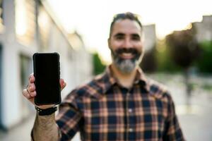 Modern businessman with beard showing blank screen of phone while standing on the city street. Focus on the phone. photo