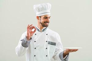 Smiling chef is holding plate and showing ok sign on gray background photo