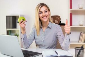 Businesswoman eating apple and showing thumb up while working in her office photo