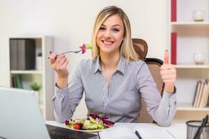 hermosa mujer de negocios disfruta comiendo ensalada y demostración pulgar arriba mientras trabajando en su oficina foto