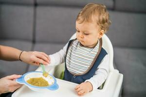 linda bebé chico aprendizaje a comer por él mismo mientras sentado en un alto silla foto