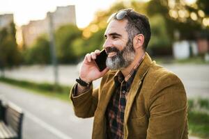 Modern businessman with beard using mobile phone while sitting on the bench in the city street photo