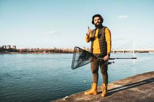 Image of fisherman with fishing tackle showing thumb up while standing by a river photo